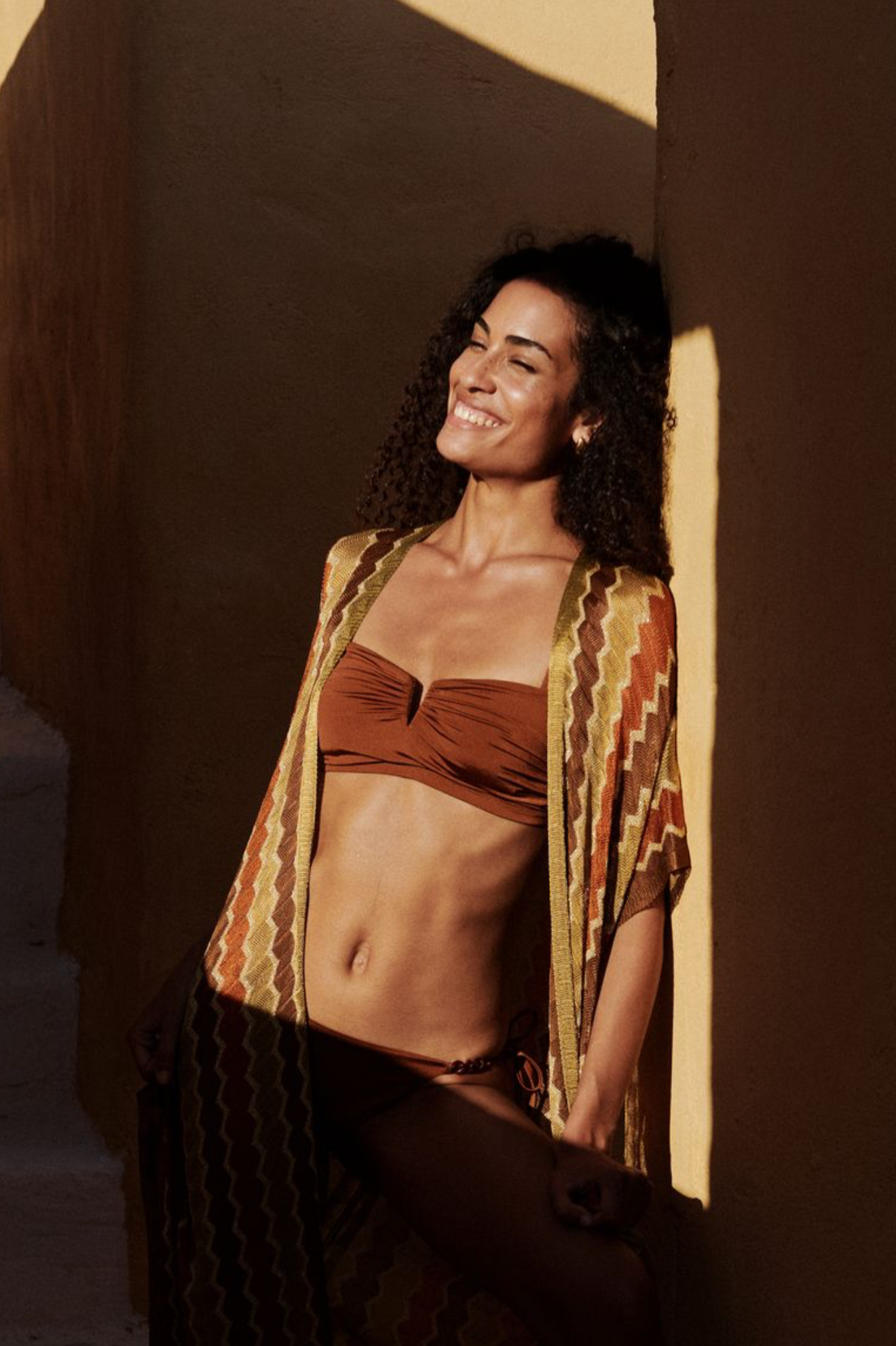 Woman in a brown bikini and patterned cover-up standing against a warm-toned wall.