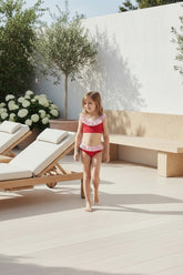 Young girl in a red swimsuit standing on a patio with lounge chairs and flowers.