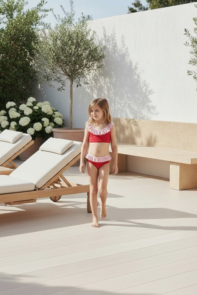 Young girl in a red swimsuit standing on a patio with lounge chairs and flowers.
