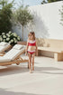 Young girl in a red swimsuit standing on a patio with lounge chairs and flowers.