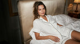 A woman reclining on a bed wearing a white short-sleeve pajama set with black piping, set in a dimly lit bedroom with a beige upholstered headboard.
