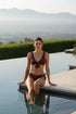 Woman in a burgundy bikini sitting by a pool with mountains in the background