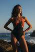 Woman in a black and red swimsuit standing on rocks by the ocean.